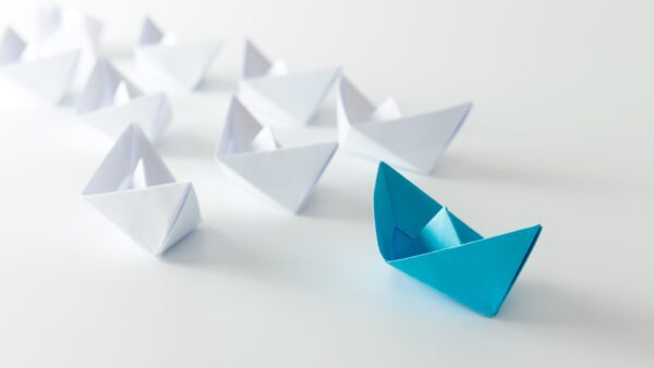 A blue paper boat is in front of many white ones.